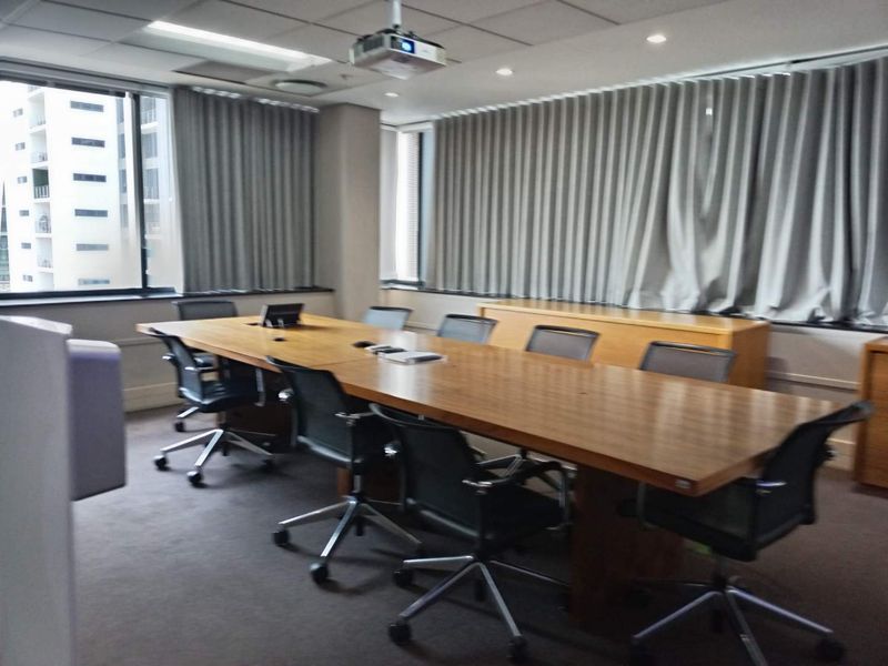 A professional boardroom featuring a large conference table, ceiling-mounted projector, recessed lighting, and expansive windows with integrated curtain tracks.