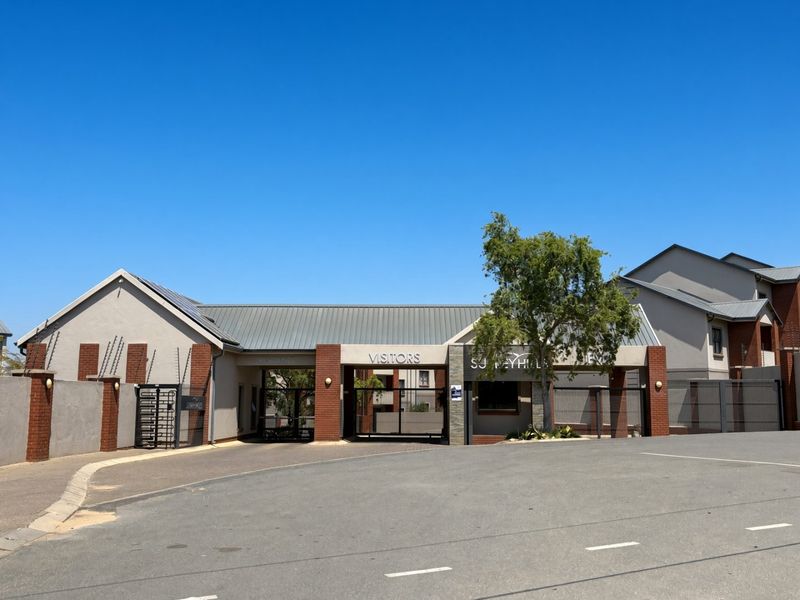 The main entrance and security gatehouse of the apartment complex, featuring brick and plaster finishes, metal roofing, and controlled access points.