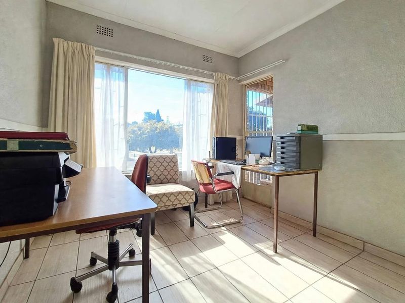 A functional home office space featuring tiled flooring, large windows providing natural light, and a security gate access point.