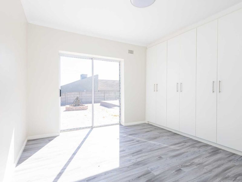 A bright, empty bedroom featuring modern laminate flooring, extensive built-in white cupboards, and a sliding glass door providing direct access to the rear garden.