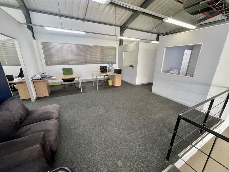 Open-plan office space featuring exposed industrial roof trusses, carpeted flooring, and internal partition walls with glass viewing panels.