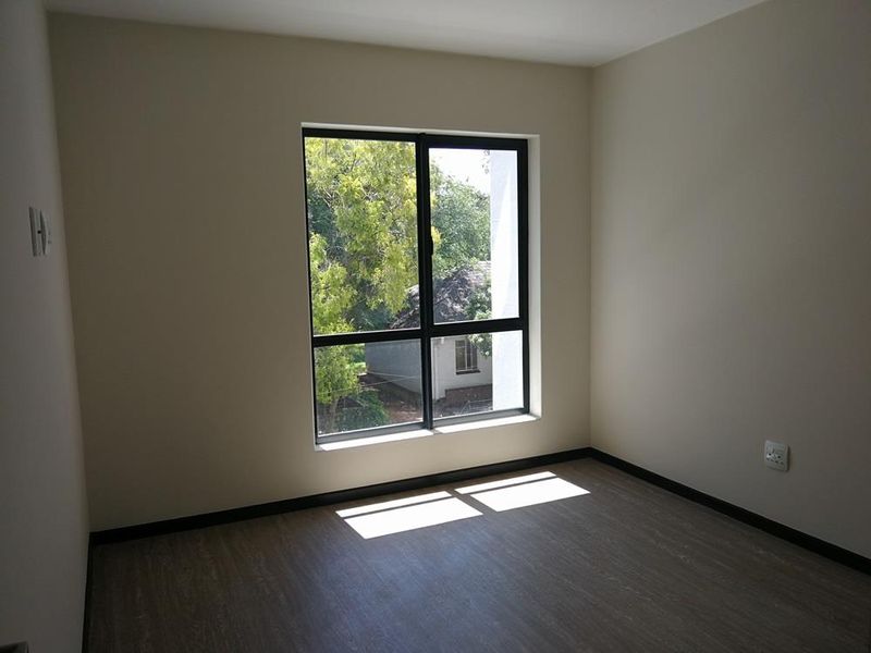 A bright, empty bedroom featuring modern laminate flooring, neutral wall finishes, and a large aluminium-framed window providing natural light.