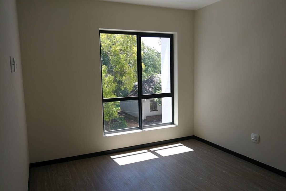 A bright, empty bedroom featuring modern laminate flooring, neutral wall finishes, and a large aluminium-framed window providing natural light.