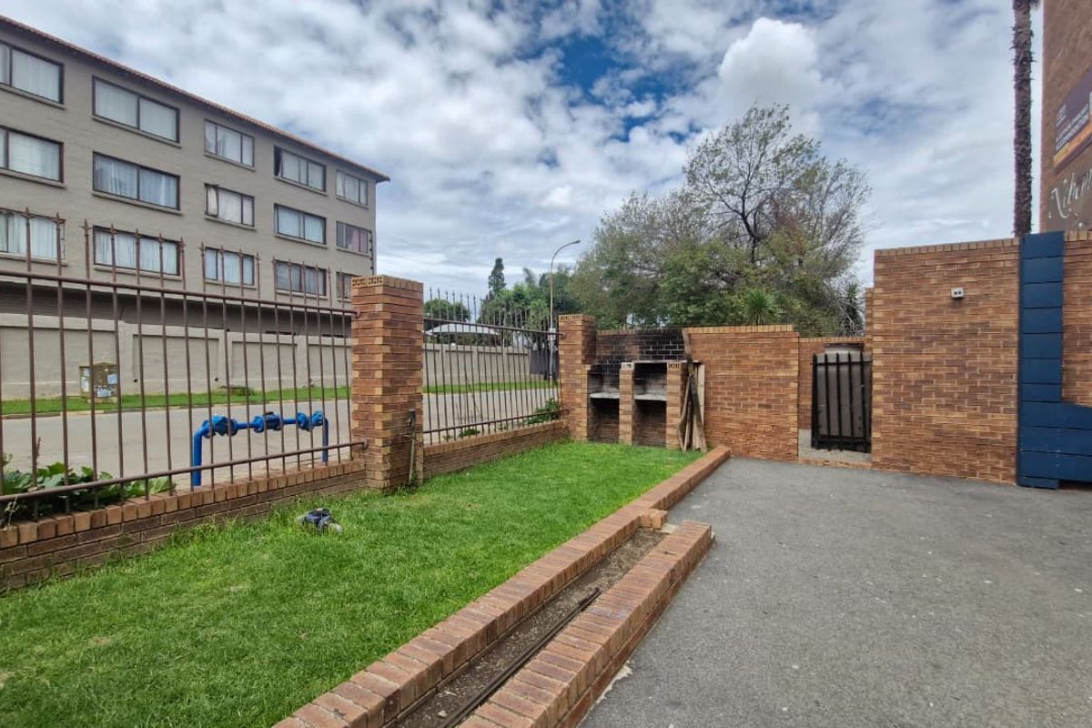 Outdoor communal area featuring a built-in brick braai, paved section, and small lawn enclosed by a metal fence.