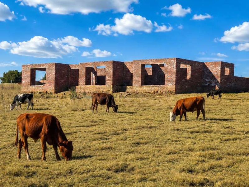 An unfinished brick structure set within a rural farm landscape, currently in the early stages of construction with open wall apertures.
