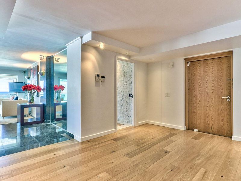 A bright, modern hallway featuring high-quality hardwood flooring, a mirrored feature wall, and recessed lighting, leading into an adjacent living area.