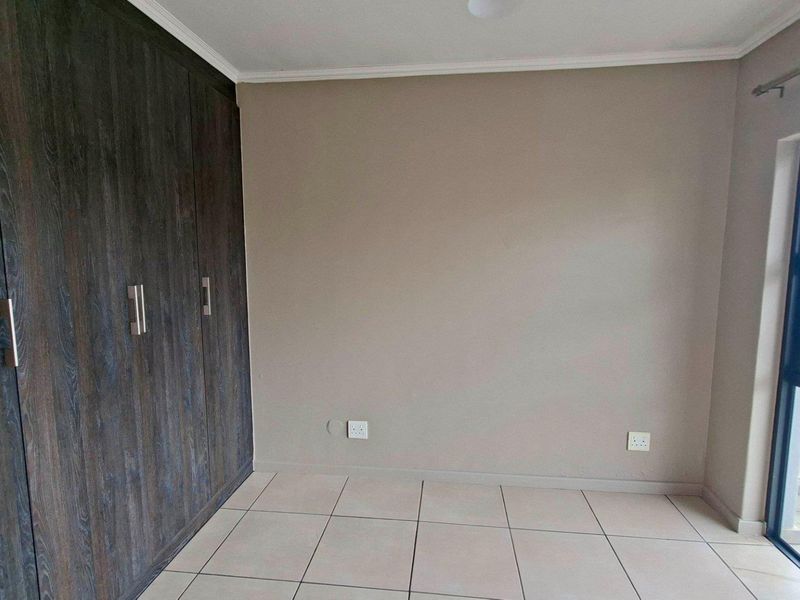 A bright, empty bedroom featuring modern dark wood built-in cupboards, neutral tiled flooring, and a sliding door leading to an outdoor area.