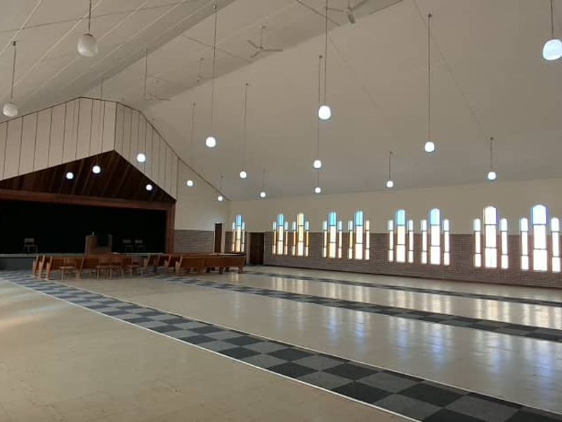 Spacious hall featuring a vaulted ceiling, arched windows, pendant lighting, and a raised stage area, suitable for events or community gatherings.