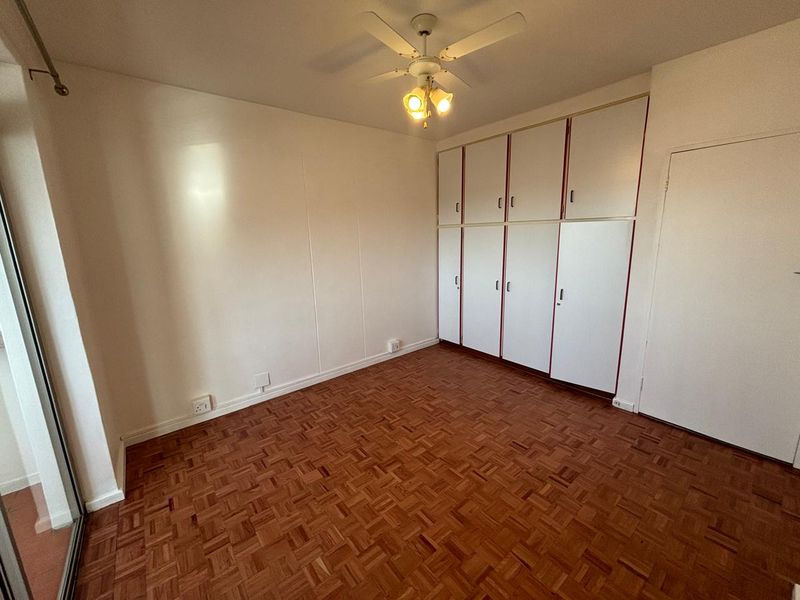 A bright, empty bedroom featuring classic parquet flooring, ample built-in cupboard storage, and a ceiling fan.