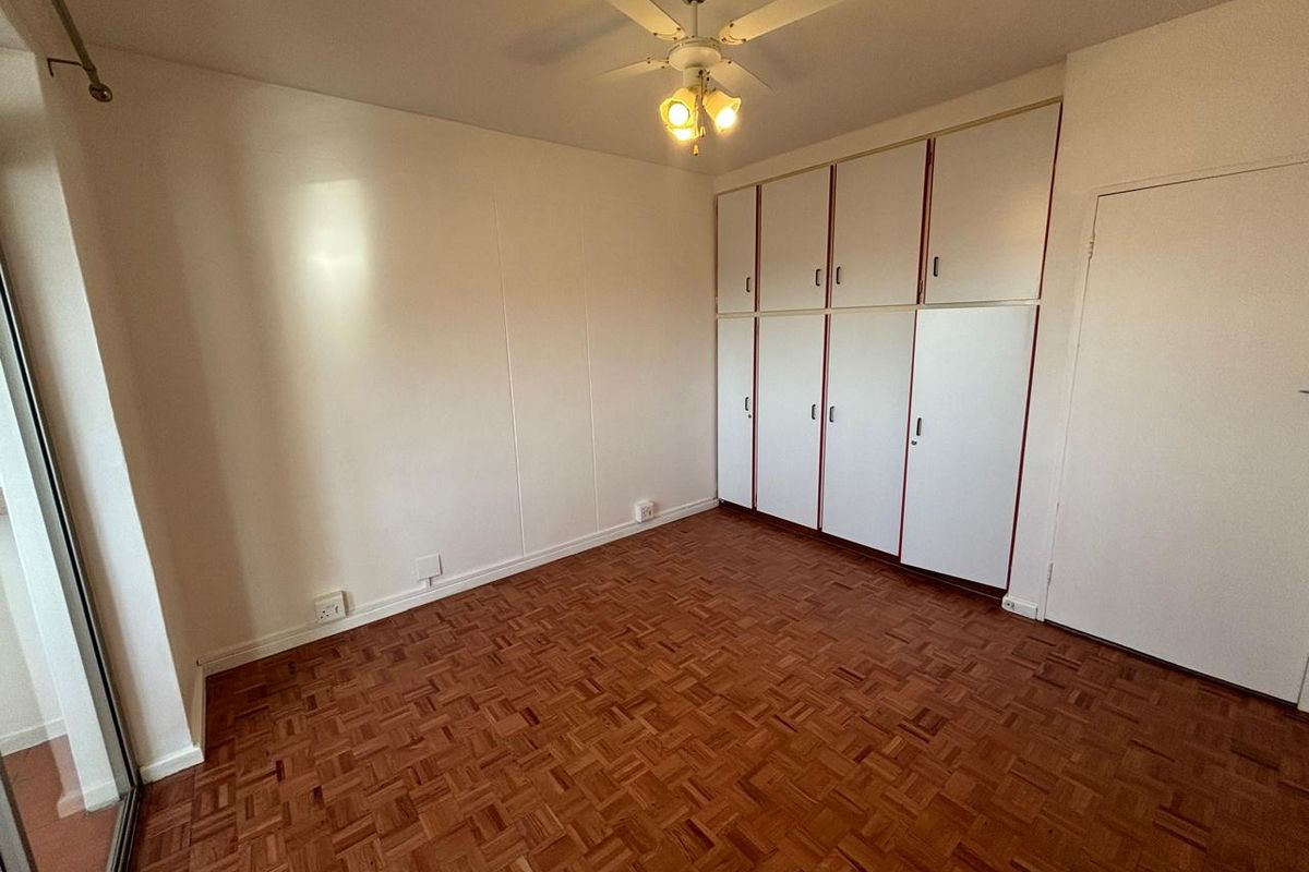 A bright, empty bedroom featuring classic parquet flooring, ample built-in cupboard storage, and a ceiling fan.