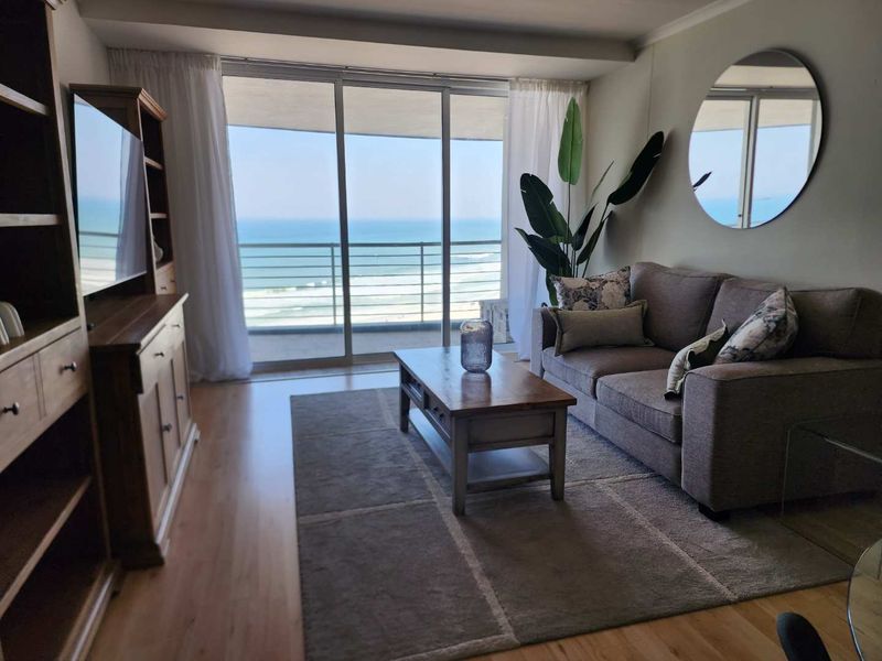 A bright living room featuring laminate flooring and large sliding glass doors that open onto a balcony with ocean views.