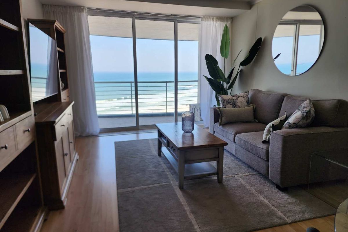 A bright living room featuring laminate flooring and large sliding glass doors that open onto a balcony with ocean views.