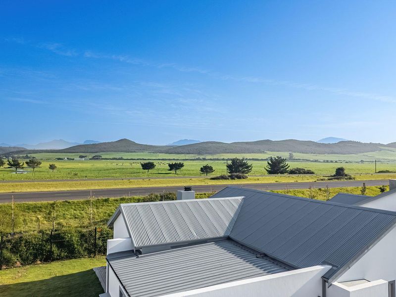 Elevated view overlooking expansive green fields and distant mountains, featuring a modern home with a corrugated metal roof in the foreground.