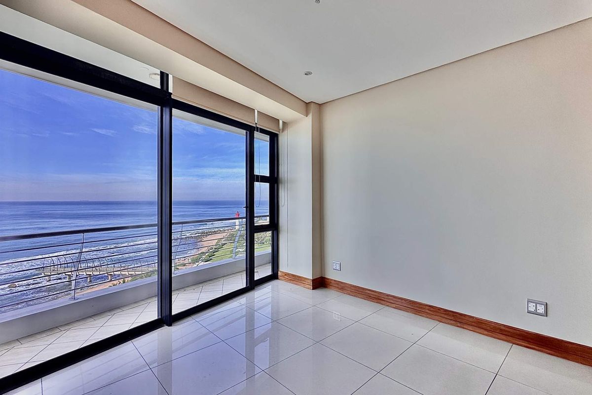 Bright bedroom featuring large floor-to-ceiling sliding glass doors that open onto a balcony with expansive ocean views and tiled flooring.