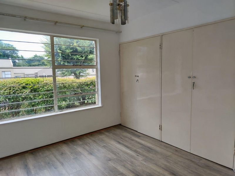 A bright, empty bedroom featuring laminate flooring, built-in cupboards, and large windows with security bars overlooking the garden.