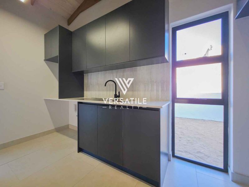 A modern, empty scullery featuring dark grey built-in cabinetry, stone countertops, a tiled backsplash, and a glass panel door leading to the exterior.