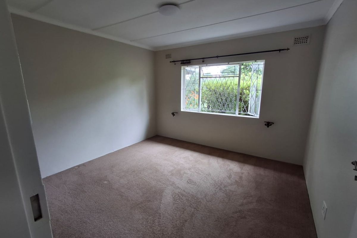 A neutral-toned bedroom featuring carpeted flooring, a window with security bars, and a simple ceiling finish.
