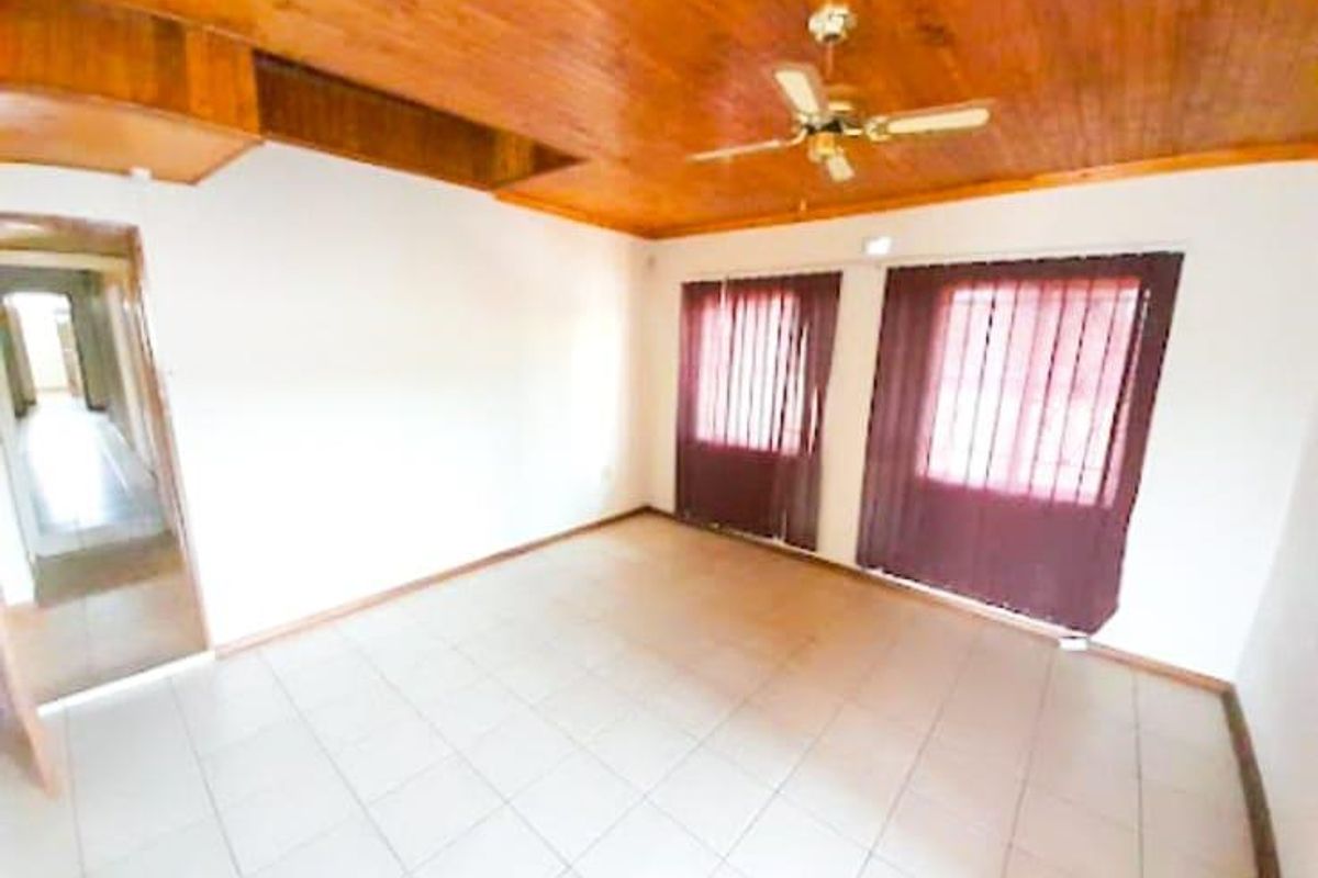 A spacious, empty living room featuring tiled flooring, a wooden panelled ceiling with a ceiling fan, and large windows with vertical blinds.
