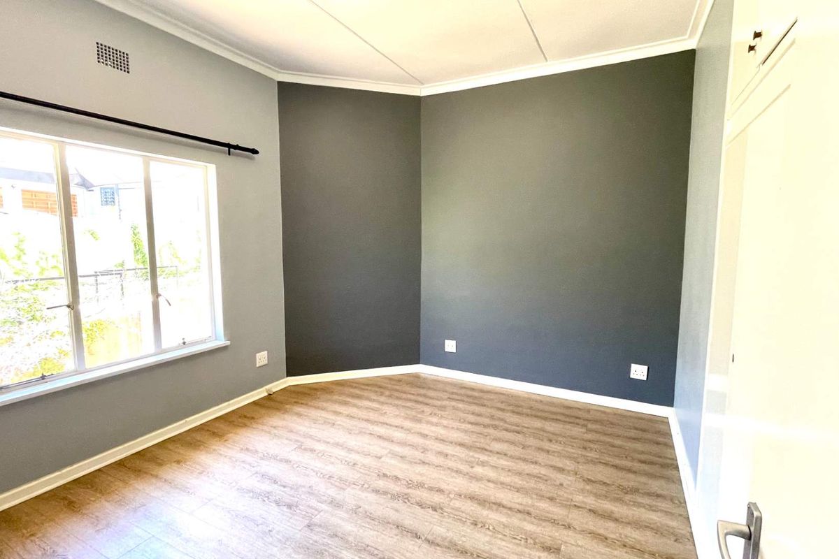 A bright, empty bedroom featuring modern wood-look laminate flooring, neutral grey walls, and a large window providing natural light.