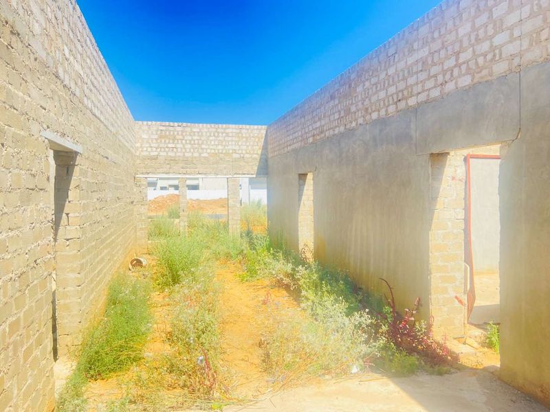An unfinished construction site showing exposed brick walls and concrete lintels with no roof or flooring, currently overgrown with vegetation.