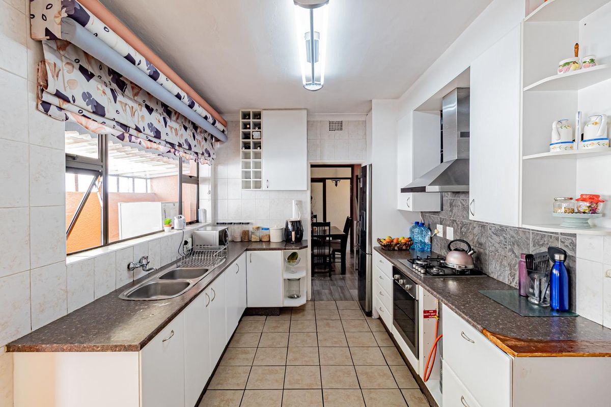 A functional galley-style kitchen featuring white cabinetry, dark laminate countertops, a built-in oven, gas hob, stainless steel extractor fan, and double basin sink.