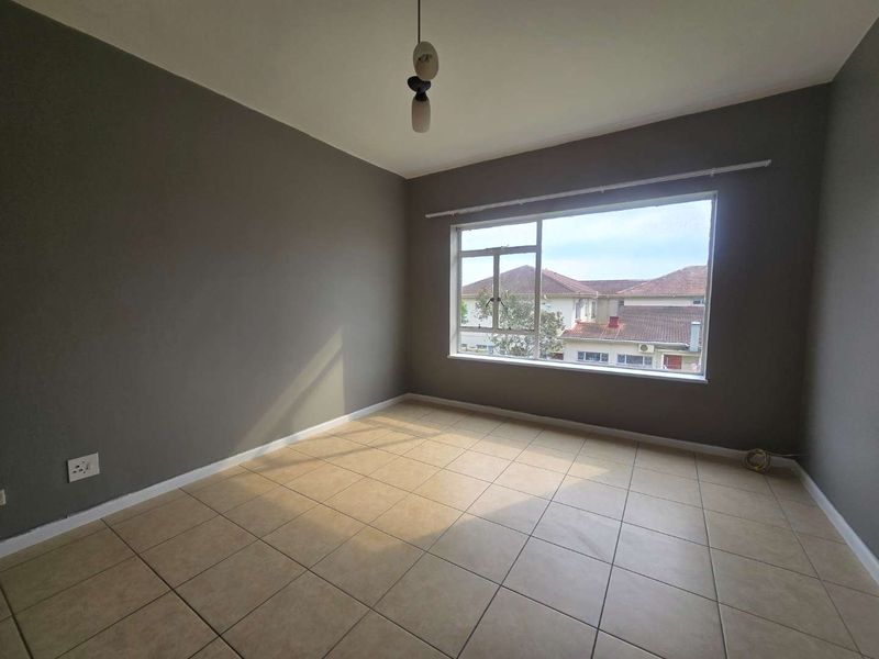 A bright, empty bedroom featuring neutral grey walls, tiled flooring, and a large window providing natural light and a view of the surrounding neighborhood.