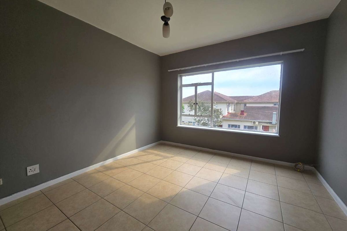A bright, empty bedroom featuring neutral grey walls, tiled flooring, and a large window providing natural light and a view of the surrounding neighborhood.