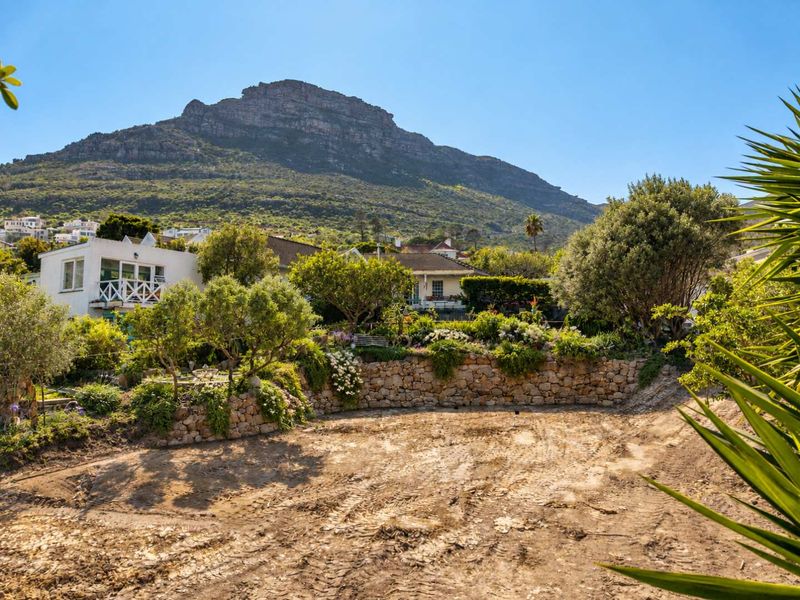 A vacant residential plot featuring a stone retaining wall, established garden beds, and mature trees with scenic mountain views.