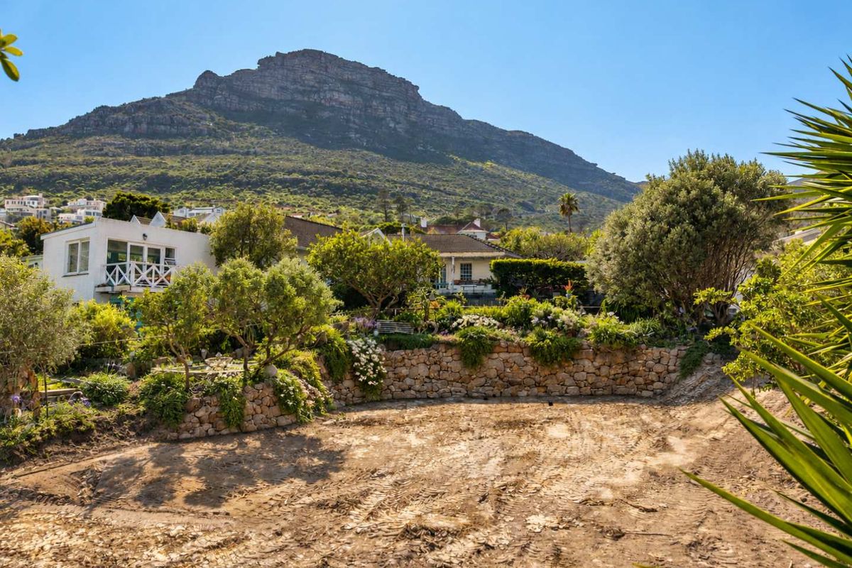 A vacant residential plot featuring a stone retaining wall, established garden beds, and mature trees with scenic mountain views.