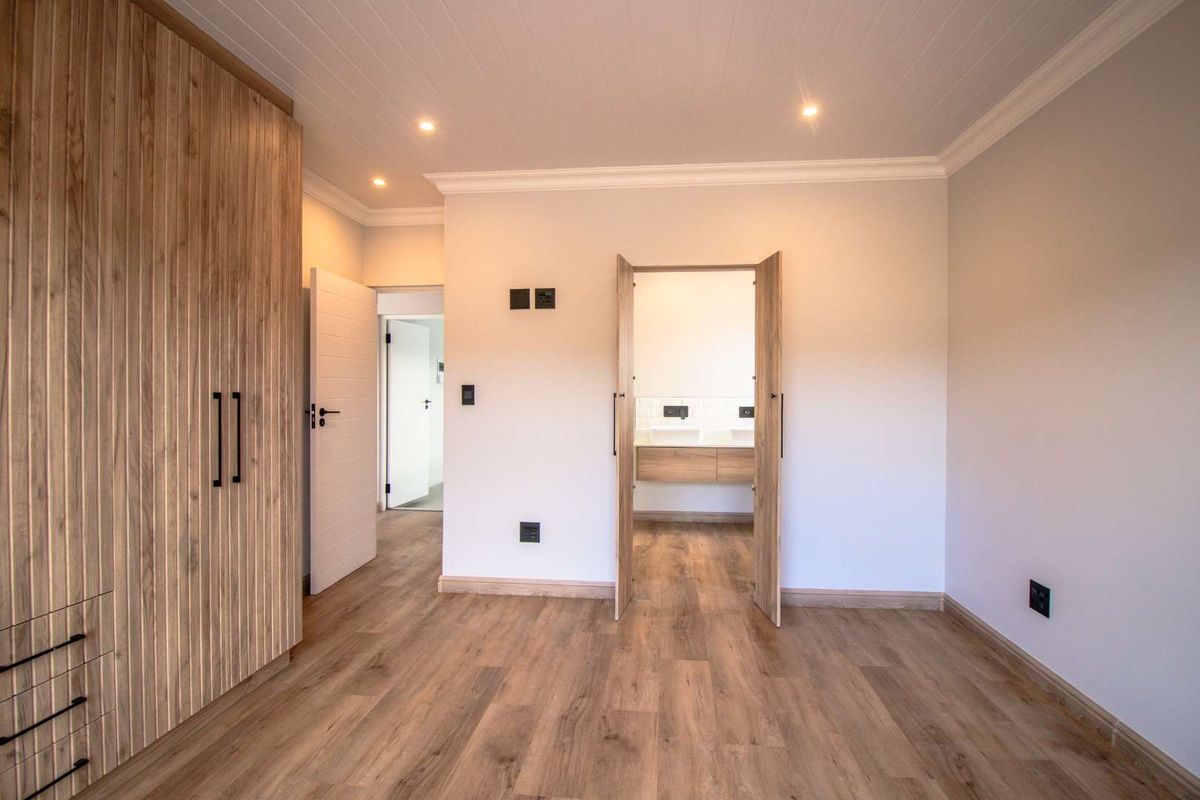 A modern, well-finished bedroom featuring light wood-look laminate flooring, built-in cabinetry, and recessed lighting, with an adjoining en-suite bathroom visible through the doorway.