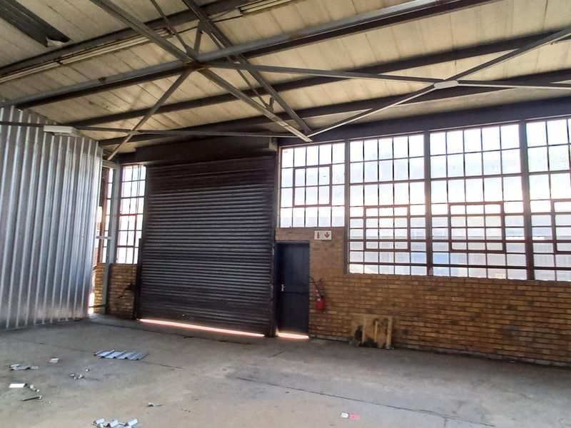 Spacious industrial utility area featuring high ceilings with exposed steel trusses, large steel-framed windows, a roller shutter door, and durable concrete flooring.