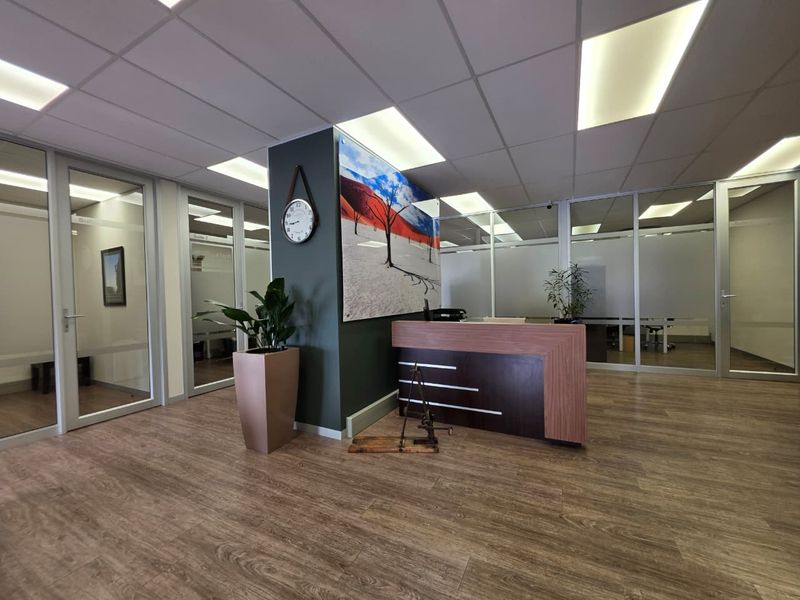 Professional office reception area featuring a modern reception desk, glass-partitioned office suites, suspended ceiling with recessed lighting, and wood-look laminate flooring.