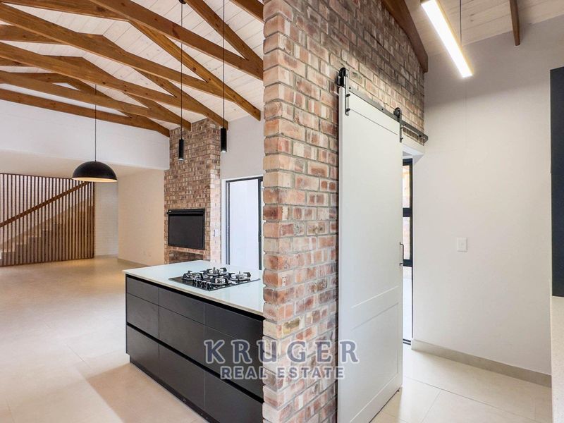 Spacious open-plan living area featuring exposed timber beams, a brick feature wall with fireplace, modern kitchen island with gas hob, and stylish barn door.