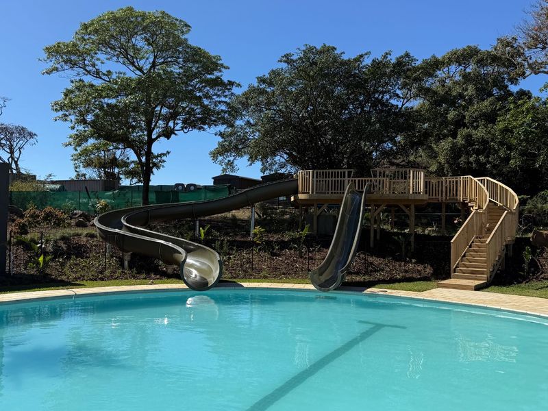 A well-maintained swimming pool area featuring a large pool, integrated water slides, and a wooden deck structure set within a landscaped garden.
