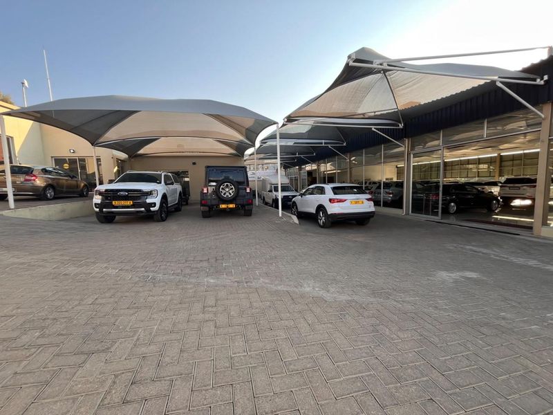 Front exterior view of a retail car dealership featuring a paved parking area, shade cloth carports, and large glass storefront windows.