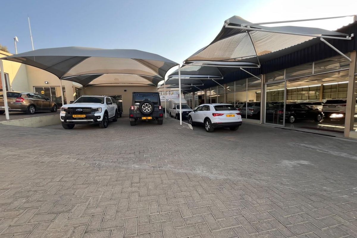 Front exterior view of a retail car dealership featuring a paved parking area, shade cloth carports, and large glass storefront windows.