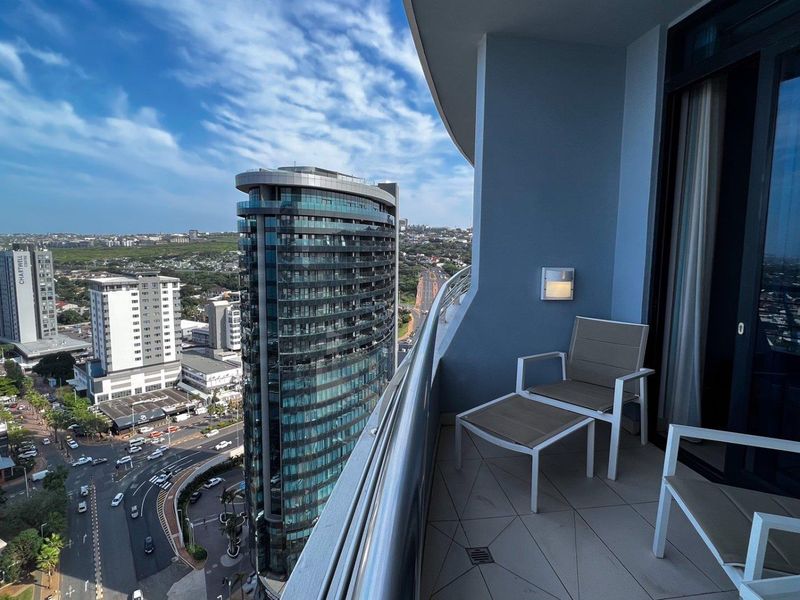 A modern apartment balcony featuring tiled flooring, a glass balustrade, and expansive urban city views.