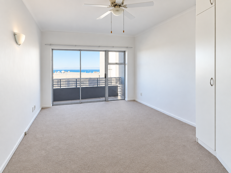 A bright, empty bedroom featuring neutral carpeting, a built-in wardrobe, a ceiling fan, and a sliding glass door leading to a balcony with ocean views.
