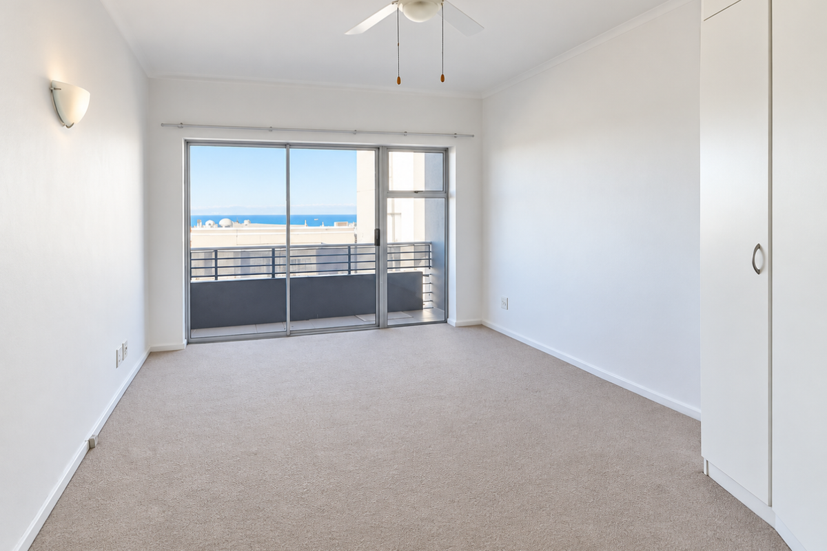 A bright, empty bedroom featuring neutral carpeting, a built-in wardrobe, a ceiling fan, and a sliding glass door leading to a balcony with ocean views.