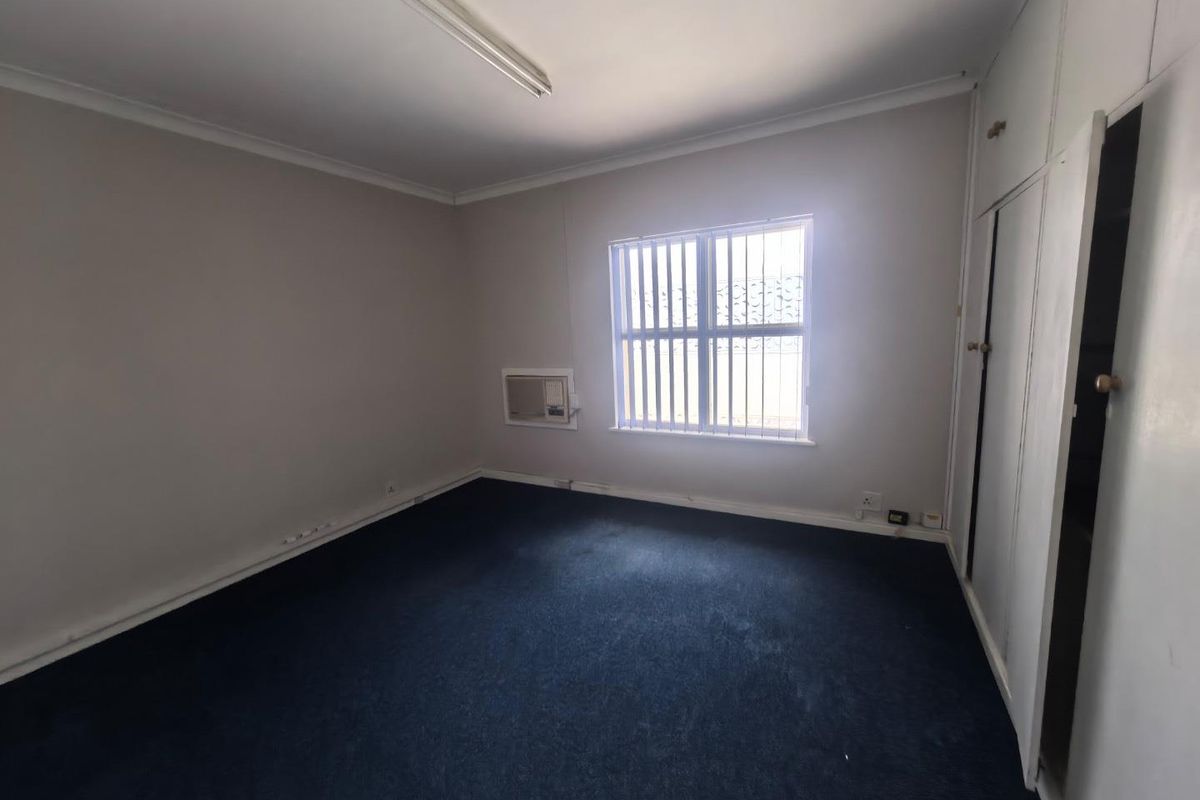 An empty bedroom featuring built-in cupboards, a wall-mounted air conditioning unit, and window security bars, finished with dark carpet flooring.