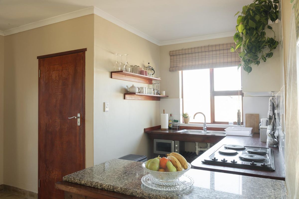 A functional kitchen featuring granite countertops, a built-in hob, stainless steel sink, and wooden shelving, offering a practical layout with natural light.
