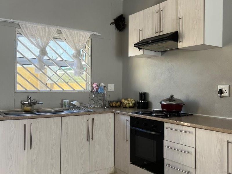 Functional kitchen featuring light wood-grain cabinetry, built-in oven and hob, stainless steel sink, and tiled flooring with security bars on the window.