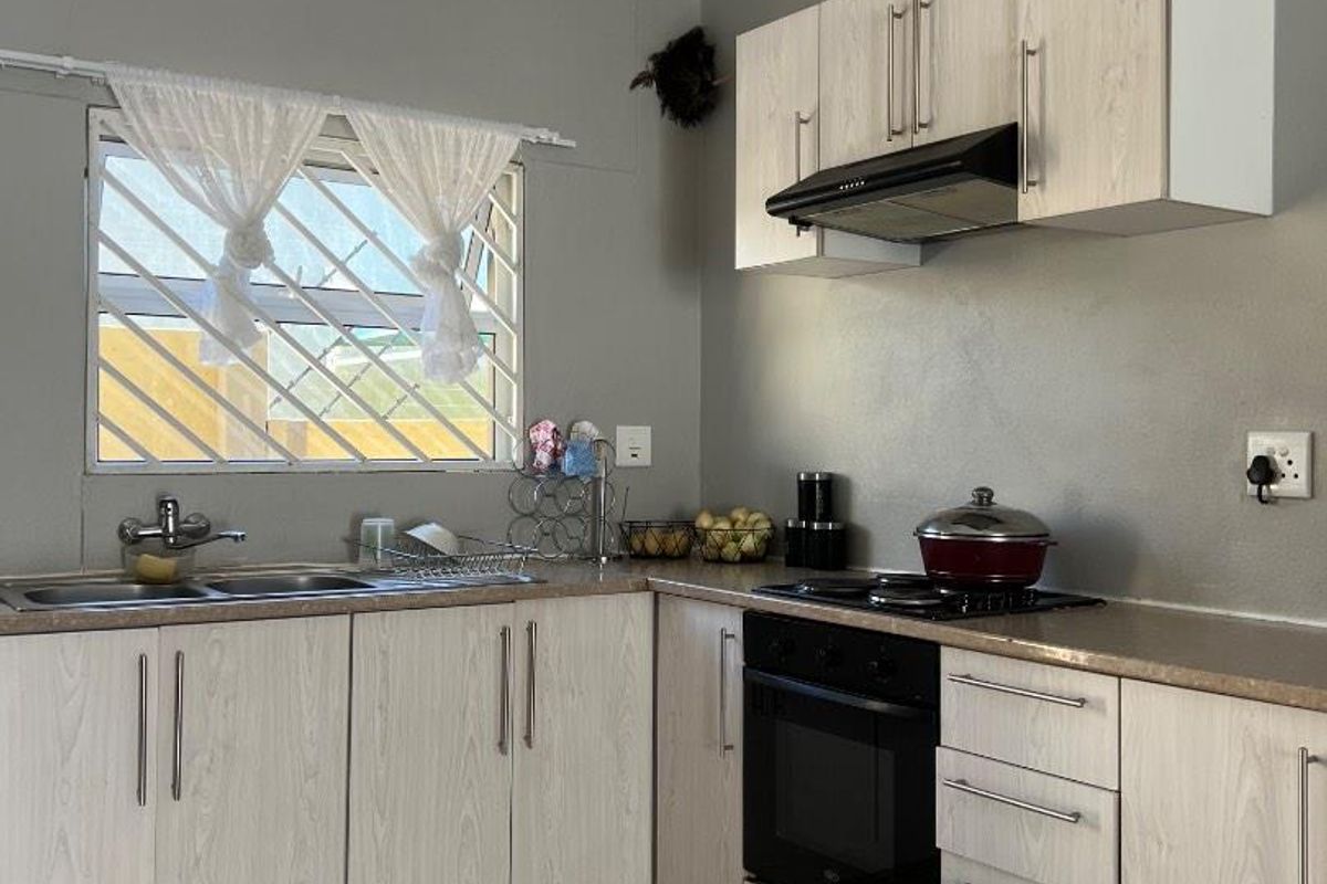 Functional kitchen featuring light wood-grain cabinetry, built-in oven and hob, stainless steel sink, and tiled flooring with security bars on the window.