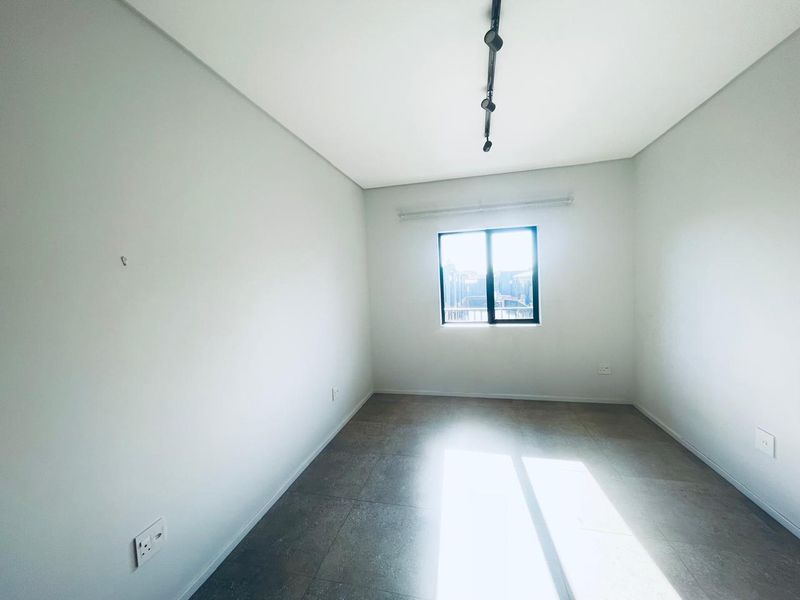 A bright, empty bedroom featuring neutral tiled flooring, modern track lighting, and a window allowing for natural light.