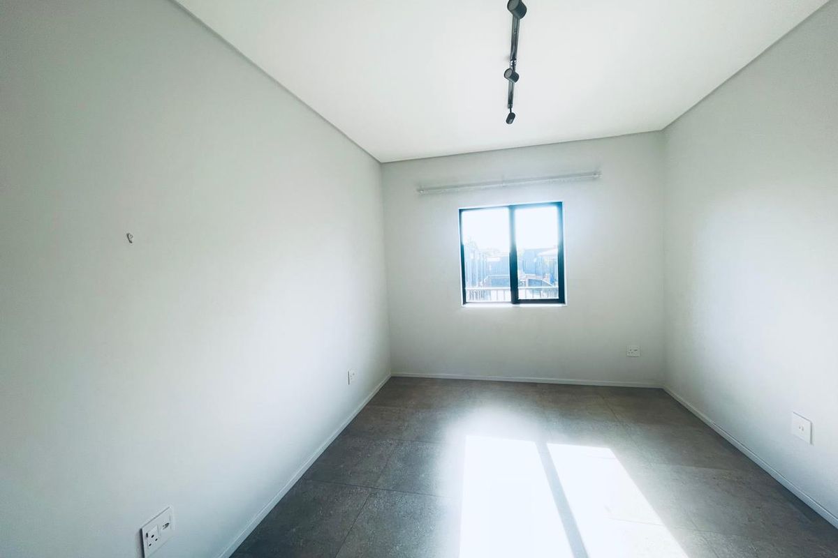 A bright, empty bedroom featuring neutral tiled flooring, modern track lighting, and a window allowing for natural light.