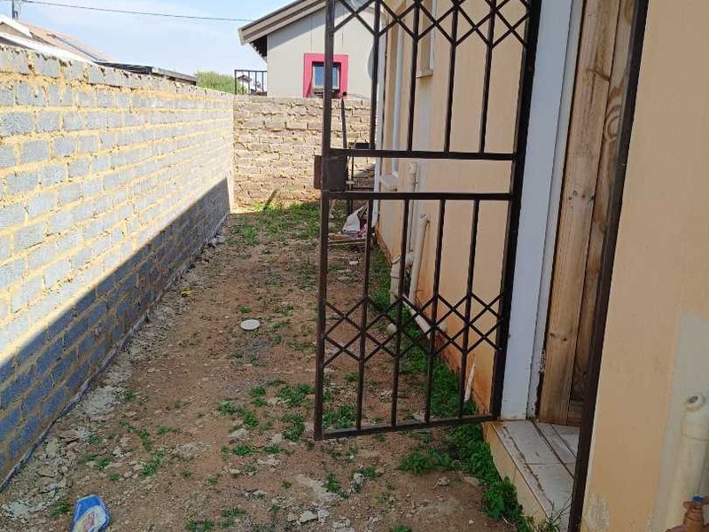 Rear exterior area featuring a security gate, brick boundary wall, and basic utility fixtures including an external tap and concrete basin.