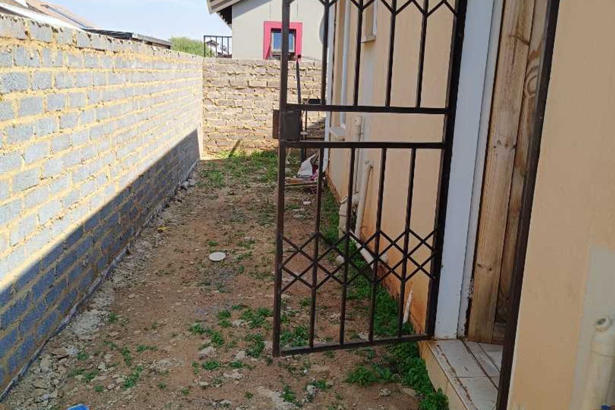 Rear exterior area featuring a security gate, brick boundary wall, and basic utility fixtures including an external tap and concrete basin.