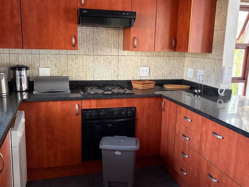 Functional kitchen featuring warm-toned wooden cabinetry, dark granite countertops, integrated electric hob, oven, and extractor fan with a neutral tiled backsplash.