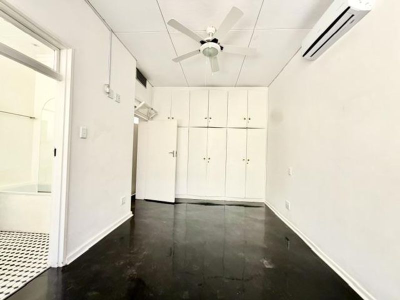 An empty bedroom featuring built-in cupboards, a ceiling fan, and a wall-mounted air conditioning unit, with direct access to an adjacent bathroom.