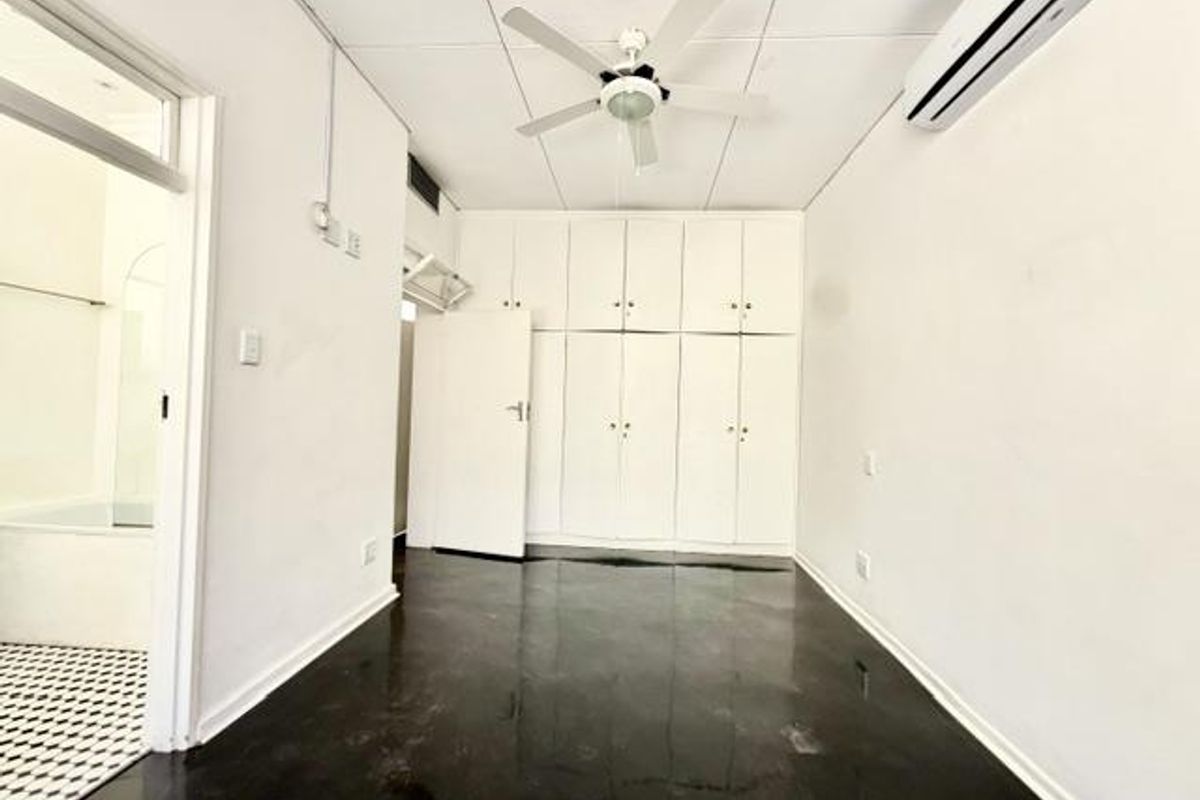 An empty bedroom featuring built-in cupboards, a ceiling fan, and a wall-mounted air conditioning unit, with direct access to an adjacent bathroom.